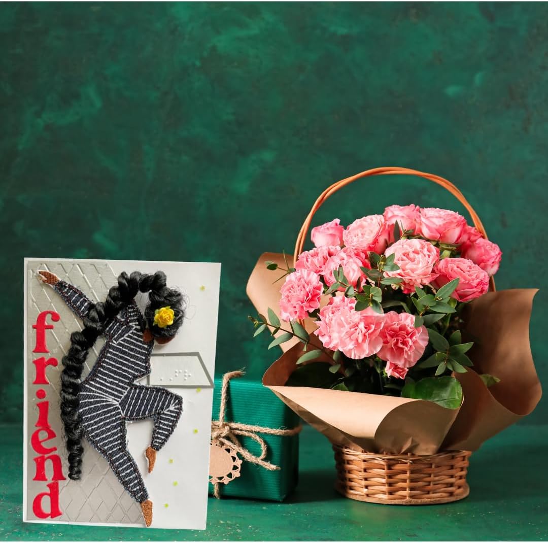 A gift arrangement featuring a pink carnation bouquet in a woven basket with brown paper wrapping, a small green gift box tied with twine, and a greeting card with a dancing figure and the word 'friend' in red.