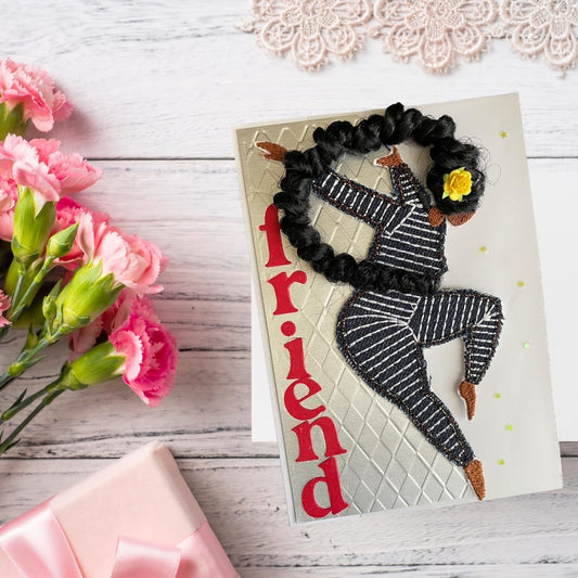 A handmade greeting card featuring a stylized figure with black braided hair, a black-and-white striped outfit, and a yellow flower, next to the word 'friend' in red. The card is styled on a white wood background with pink carnations, a pink gift box, and a lace doily.
