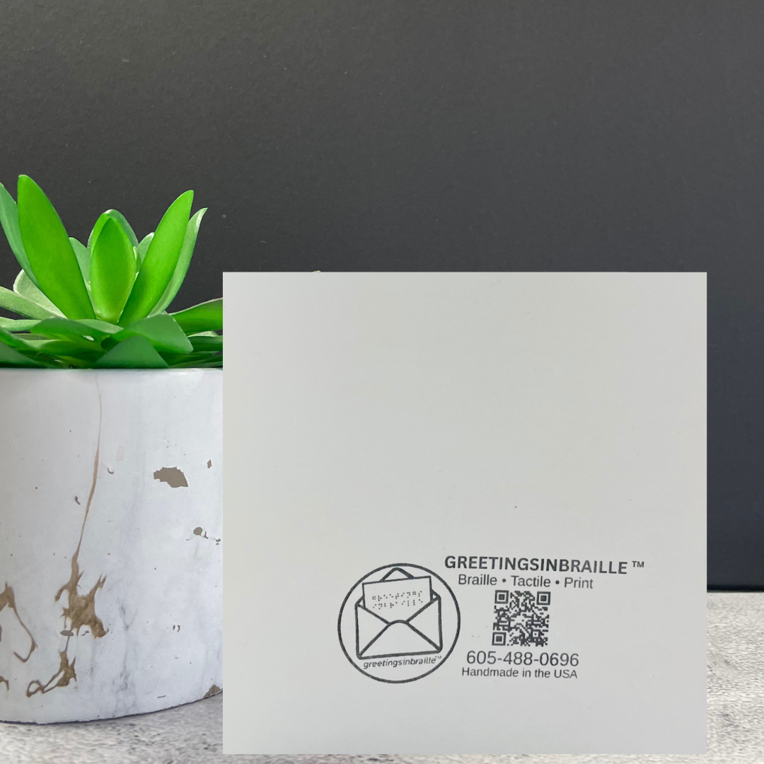 A square white tactile print card with black text and a QR code, standing next to a small green succulent in a white planter against a dark gray background.