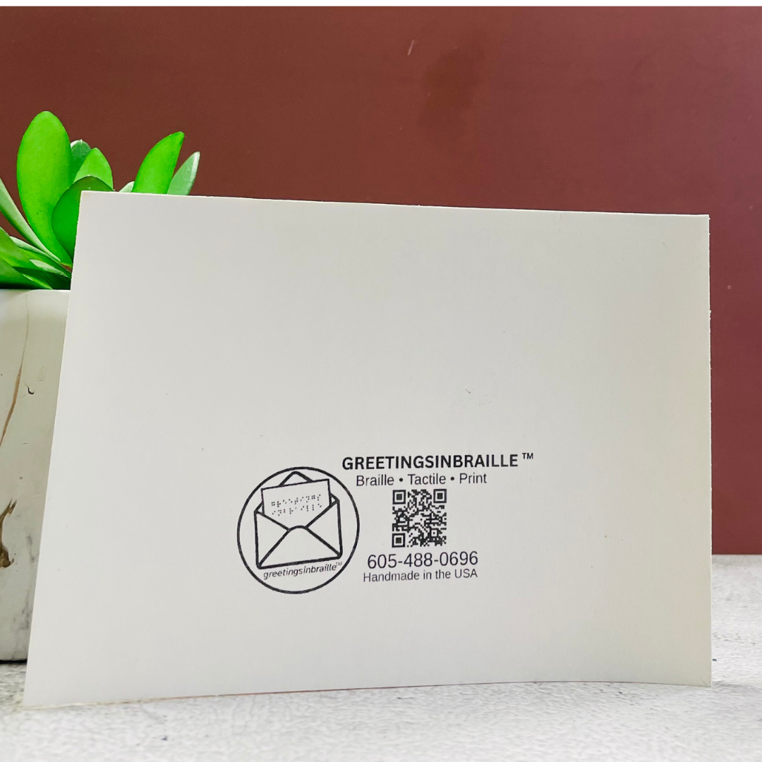 A white tactile greeting card with black printed text and a QR code, standing upright on a light surface against a dark reddish-brown background with a green plant in the corner.
