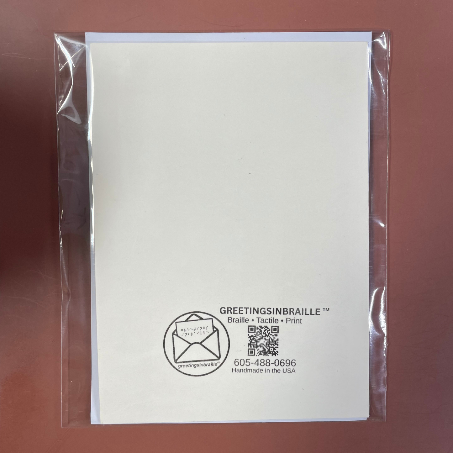 A white GreetingsinBraille tactile greeting card inside a clear plastic sleeve, displayed on a solid dark reddish-brown background.