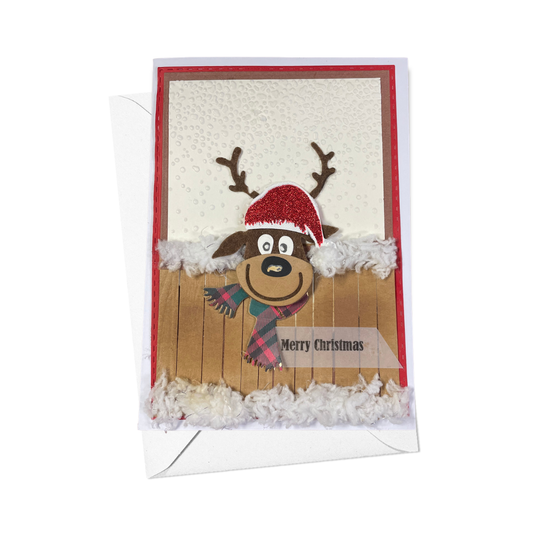 A handmade Christmas greeting card featuring a cartoon reindeer wearing a red Santa hat and plaid scarf, with a wooden fence and white fluffy trim. The card is shown with its white envelope against a plain white background.