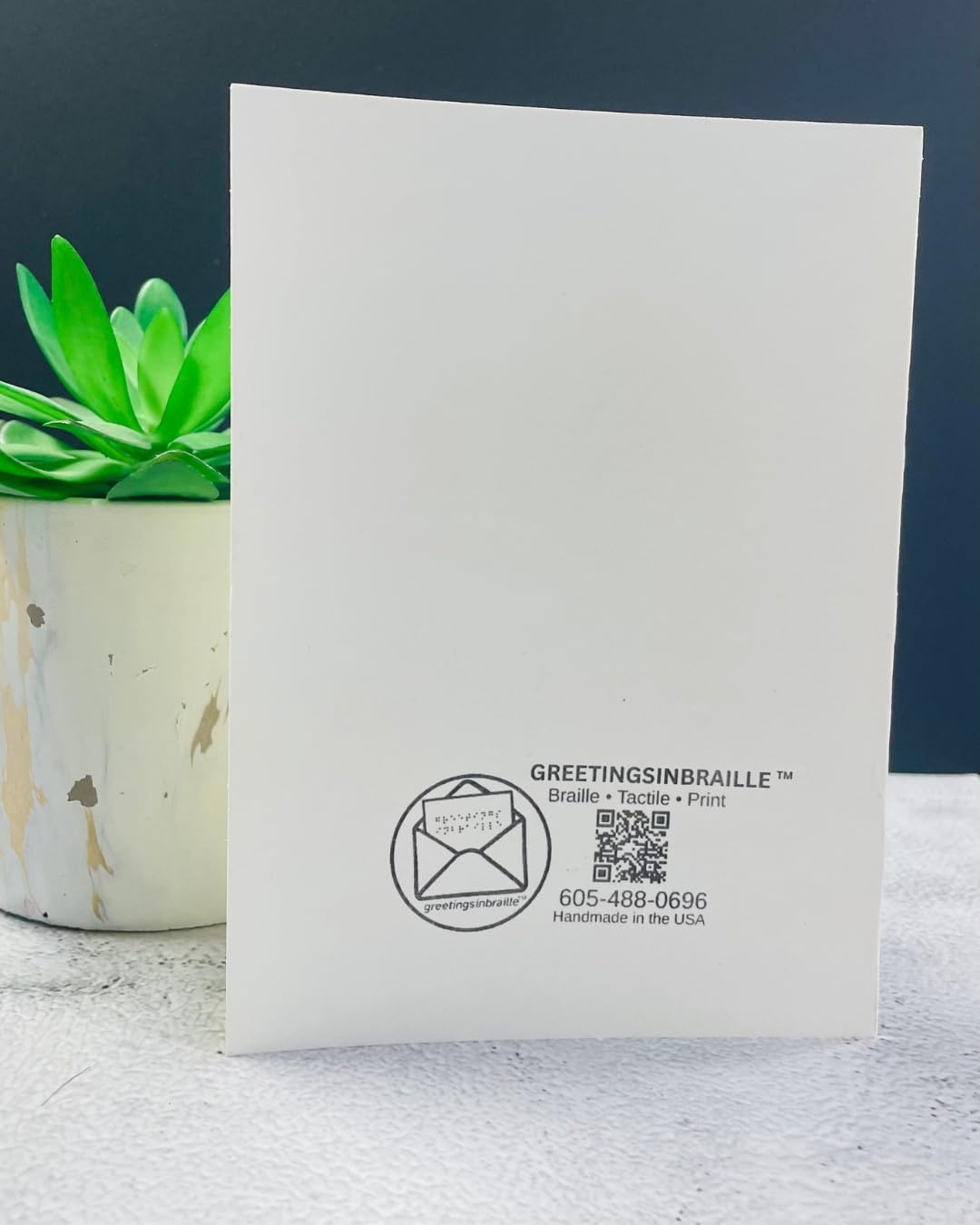 A white greeting card standing upright on a textured white surface, with a small green succulent in a beige pot to the left. The card has black printed text and a QR code near the bottom, including the brand name 'GREETINGSINBRAILLE'.