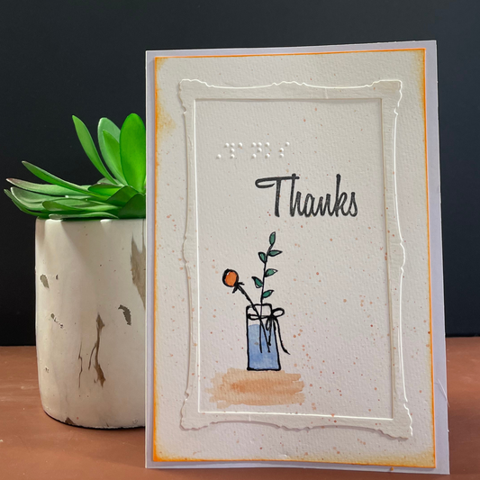 A handmade greeting card with a layered white embossed frame, the word 'Thanks' in black script, and a small illustration of a blue vase with a flower, photographed standing on a wooden surface next to a potted succulent.
