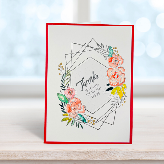 A handmade greeting card with a red border, featuring a watercolor floral wreath and the text 'Thanks SO GRATEFUL FOR ALL THAT YOU DO' in black script, standing on a light wooden surface with a soft-focus background.
