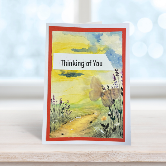 A handmade greeting card standing upright on a light wooden surface. The card features a watercolor landscape with a yellow and green field, a butterfly, wildflowers, and a red border.