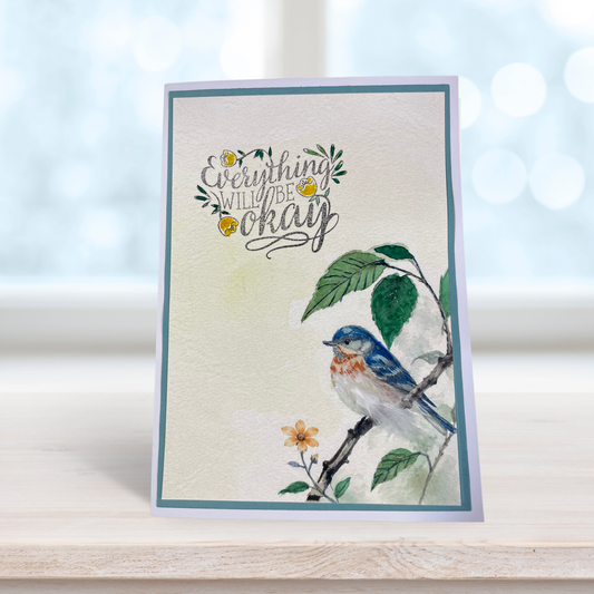 A handmade greeting card with a watercolor bluebird on a branch and the phrase 'Everything will be okay' in script, standing on a light wooden surface with a soft-focus bokeh background.