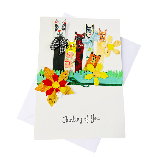 A white greeting card with a colorful handmade-style illustration of five cats standing in grass, decorated with paper flowers, and the words 'Thinking of You' printed and in braille near the bottom. A white envelope is partially visible behind the card on a white background.