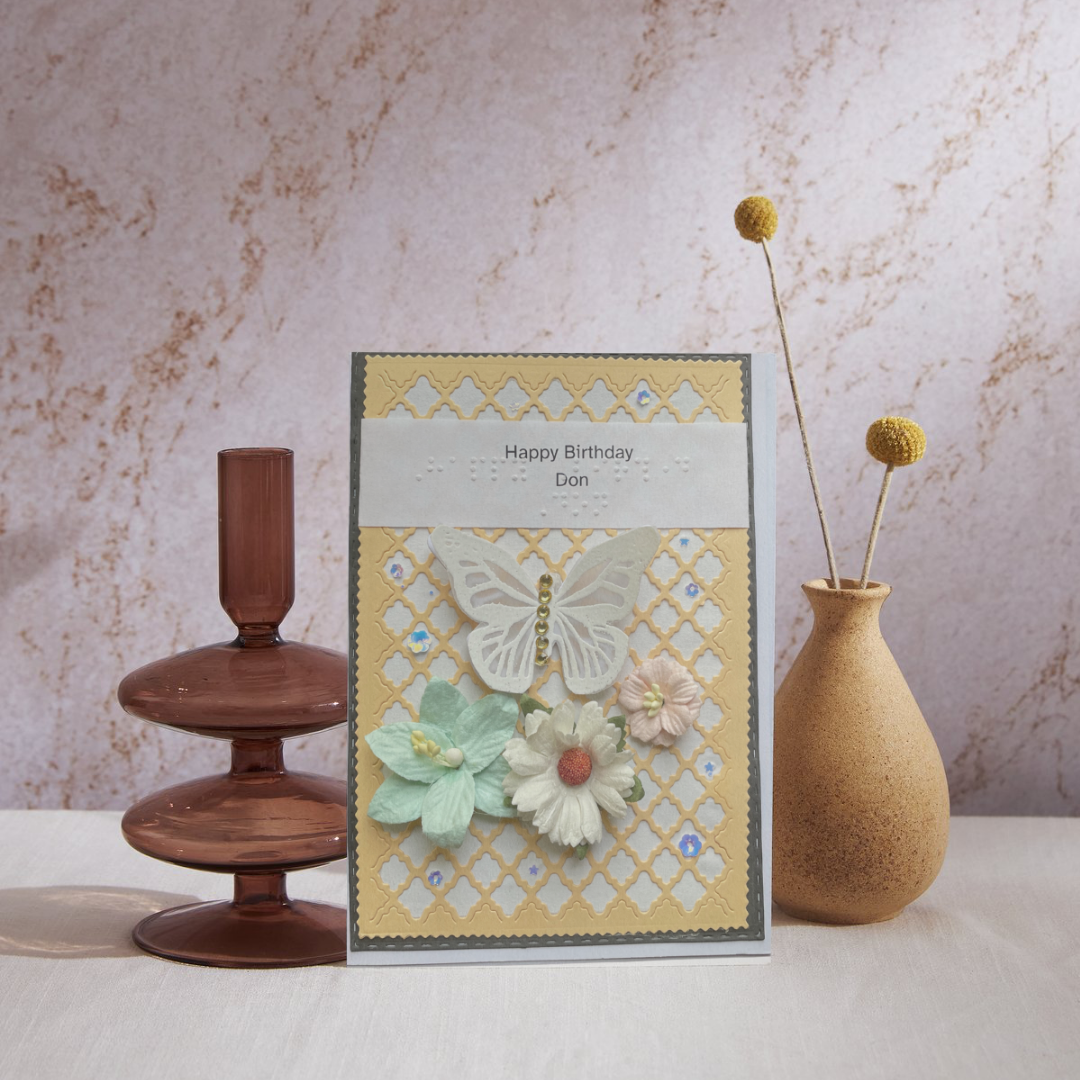 Decorative birthday card with flowers and butterfly design, placed between a wooden cake stand and a vase on a marble surface.