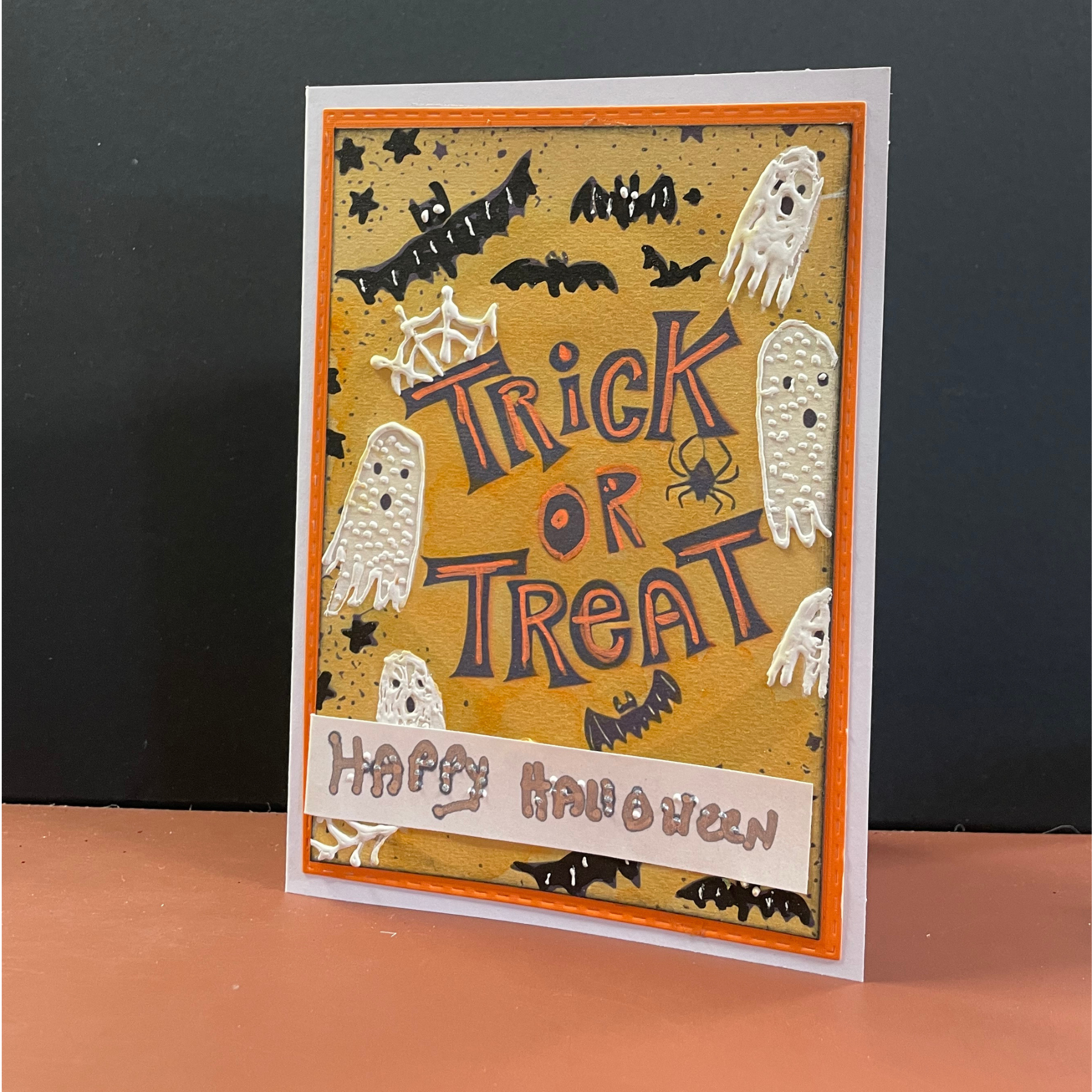 A handmade Halloween greeting card with a layered design, featuring 'Trick or Treat' in stylized lettering on a textured tan background with white ghosts, black bats, and a 'Happy Halloween' banner at the bottom. The card is displayed standing upright on a peach-colored surface against a dark gray background.