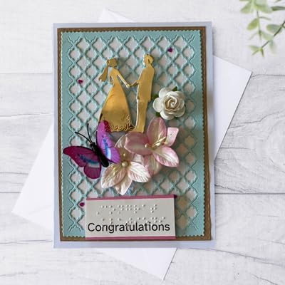 A handmade congratulations card featuring a turquoise and gold trellis background, gold bride and groom silhouettes, pink and white flowers, a purple butterfly, and a 'Congratulations' banner with braille, displayed on a white envelope over a light wood surface.
