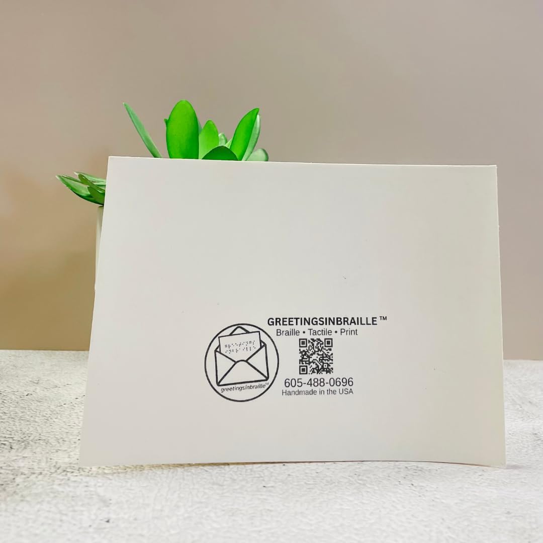 A white greeting card from GreetingsinBraille, featuring a black printed logo, QR code, and contact information, standing upright on a textured white surface with a small green plant in the background.
