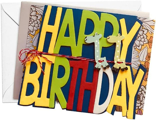 A colorful handmade-style birthday card with layered letters spelling HAPPY BIRTHDAY in yellow, green, red, and white on a dark blue background. Two small giraffe cutouts are tied across the front with red-and-white string, and a white envelope is visible behind the card.