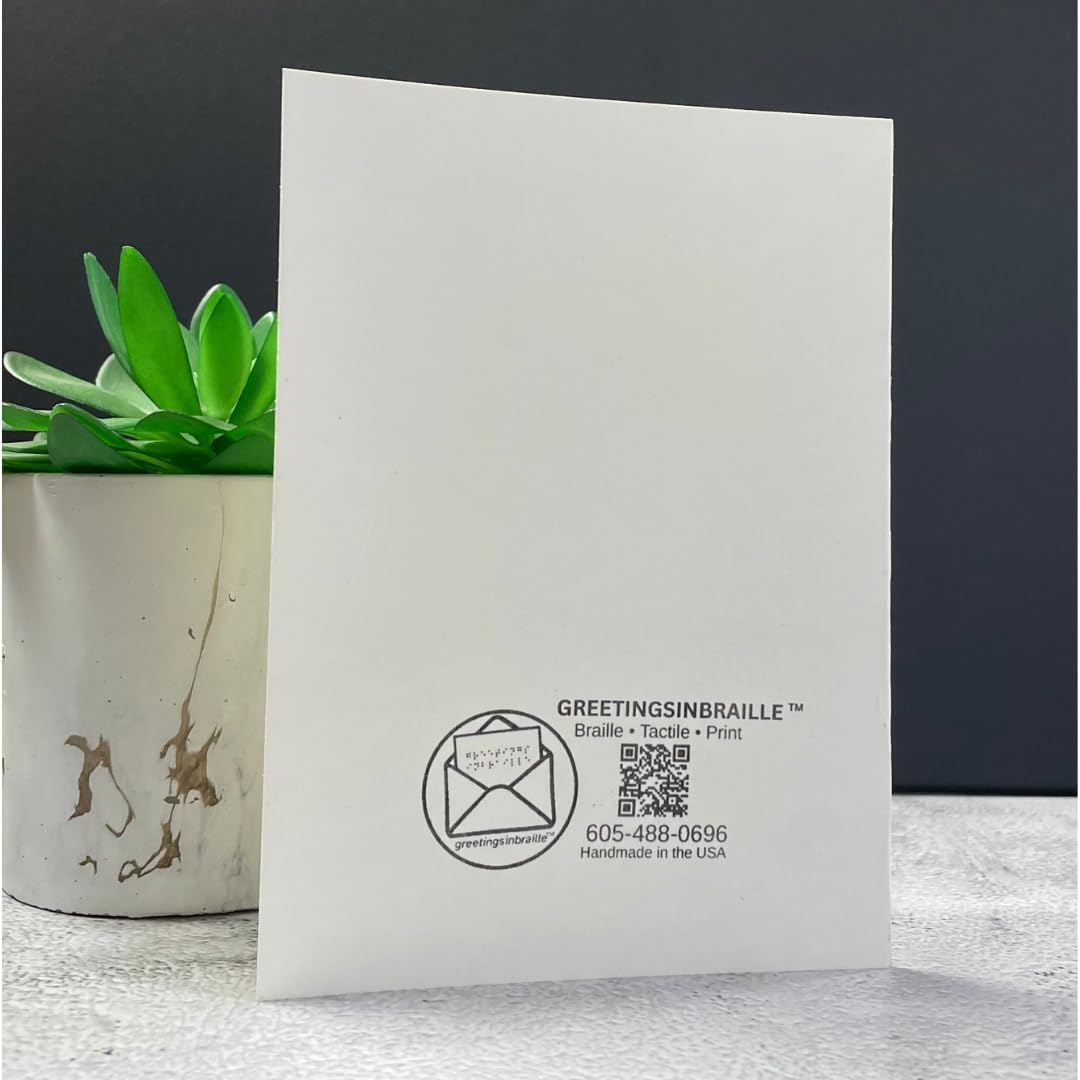 A white greeting card standing upright on a light surface, with a small green succulent in a white pot at the left and a dark gray background behind it. The card has black printed text and a QR code near the bottom, including the brand name GreetingsinBraille.