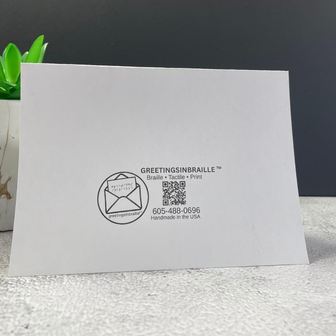 A white square card or envelope-like product standing upright on a textured white surface, with black printed text reading GREETINGSINBRAILLE and a circular logo. A small green plant in a white pot is partially visible at the left edge against a dark gray background.