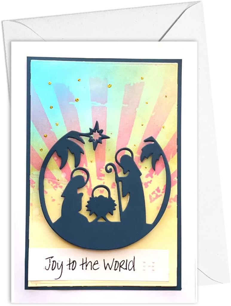 A handmade greeting card featuring a dark navy silhouette of a nativity scene against a pastel sunburst background with gold glitter accents, with the text 'Joy to the World' printed on a white strip at the bottom. The card is shown with its white envelope behind it on a white background.