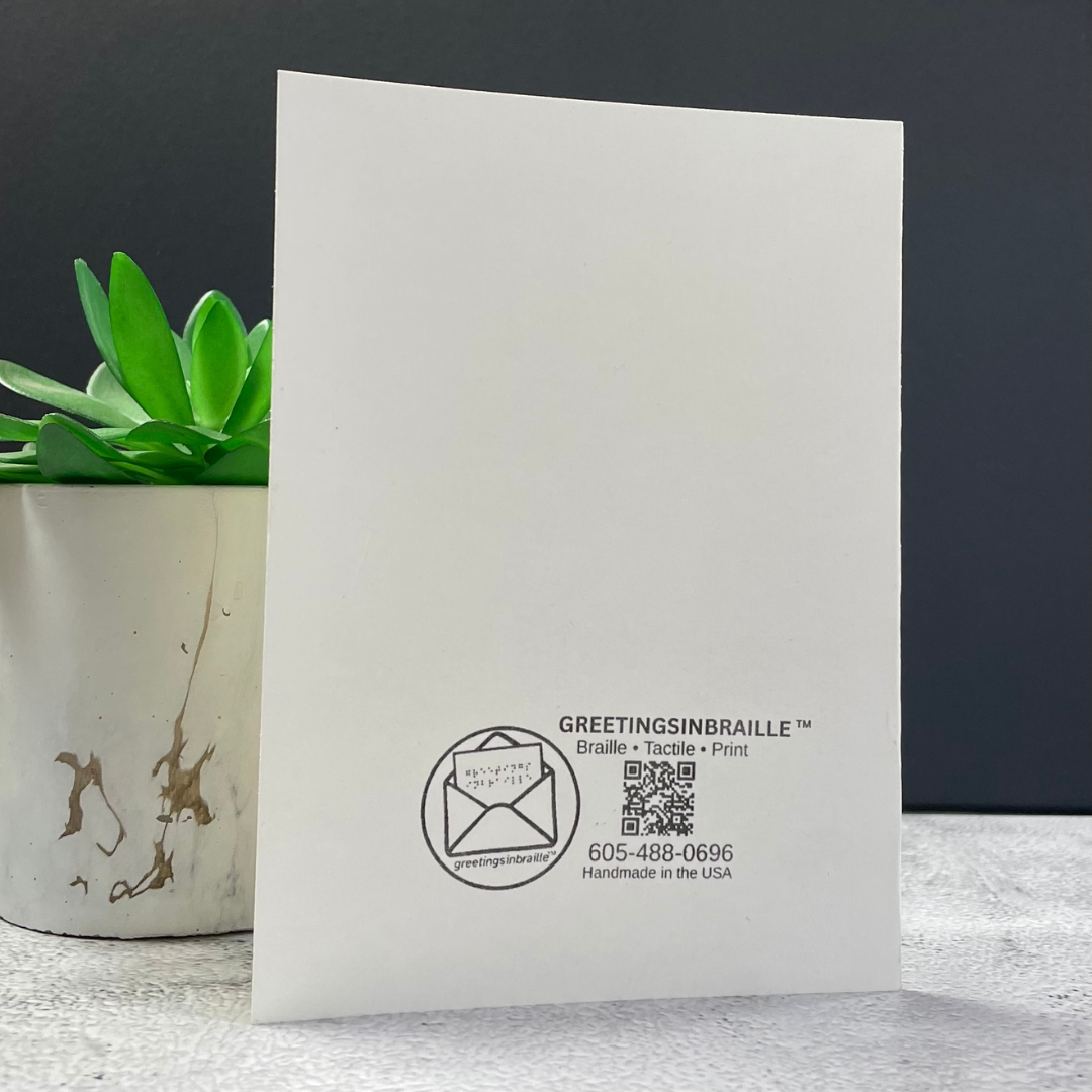 A white rectangular card or paper product standing upright on a light surface, with black printed text and logo near the bottom reading GREETINGSINBRAILLE and a QR code. A small green succulent in a white marbled pot sits to the left against a dark gray background.