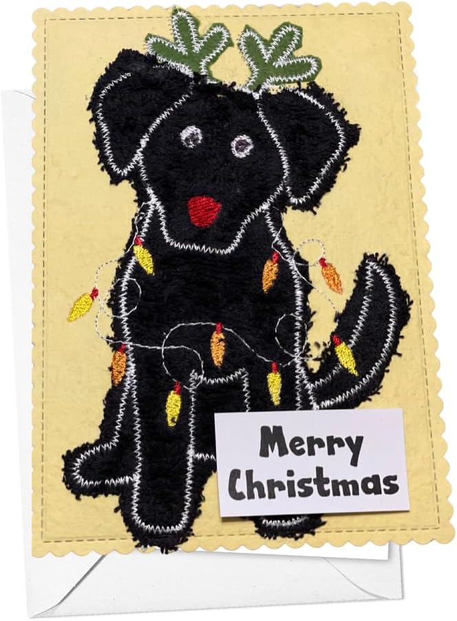 A handmade Christmas card featuring a black dog applique with green leafy antlers, white stitched outlines, a red nose, and yellow/orange Christmas lights. A small white tag on the card reads 'Merry Christmas'.