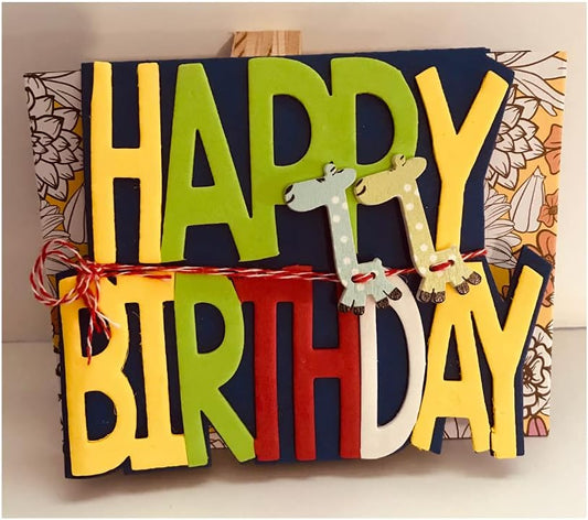 A handmade layered paper 'HAPPY BIRTHDAY' card or sign with colorful letters in yellow, green, red, and white, decorated with two small giraffe cutouts and tied with red string, shown against a patterned backing.
