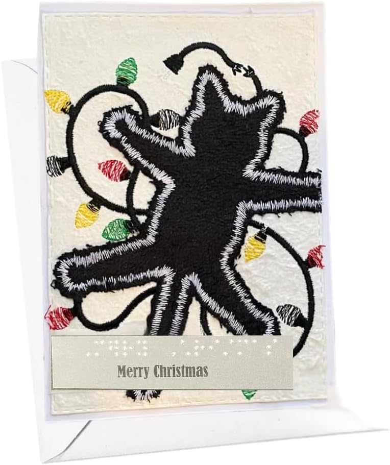 A square Christmas greeting card with a black embroidered star design outlined in white, surrounded by multicolored Christmas lights on a cream background, with a beige banner reading 'Merry Christmas' at the bottom. The card is shown standing upright with a white envelope behind it.