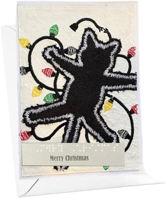 A square Christmas greeting card with a black embroidered star design outlined in white, surrounded by multicolored Christmas lights on a cream background, with a beige banner reading 'Merry Christmas' at the bottom. The card is shown standing upright with a white envelope behind it.