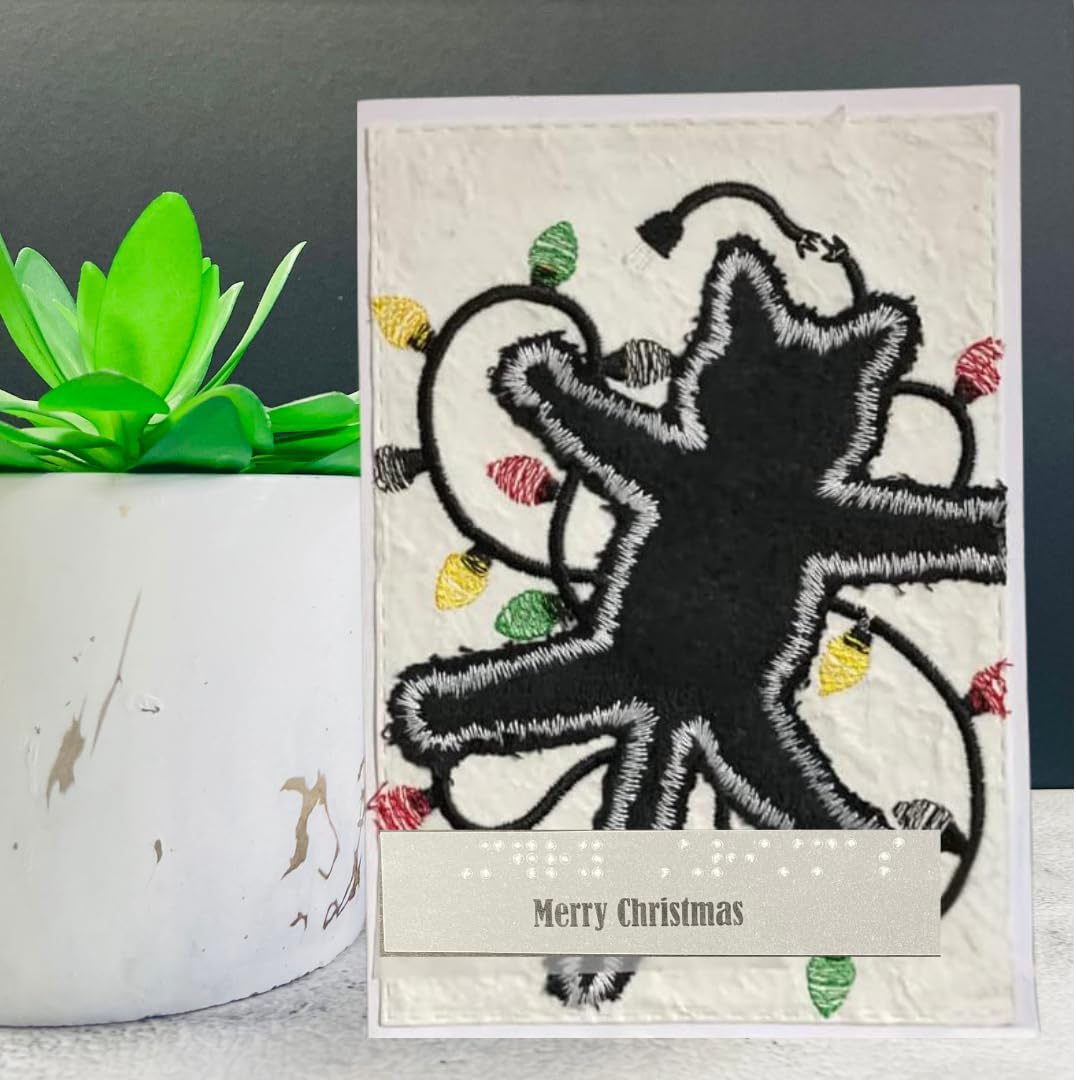 A handmade Christmas greeting card with a black star design outlined in white, surrounded by colorful thread-like Christmas lights, placed next to a potted succulent.