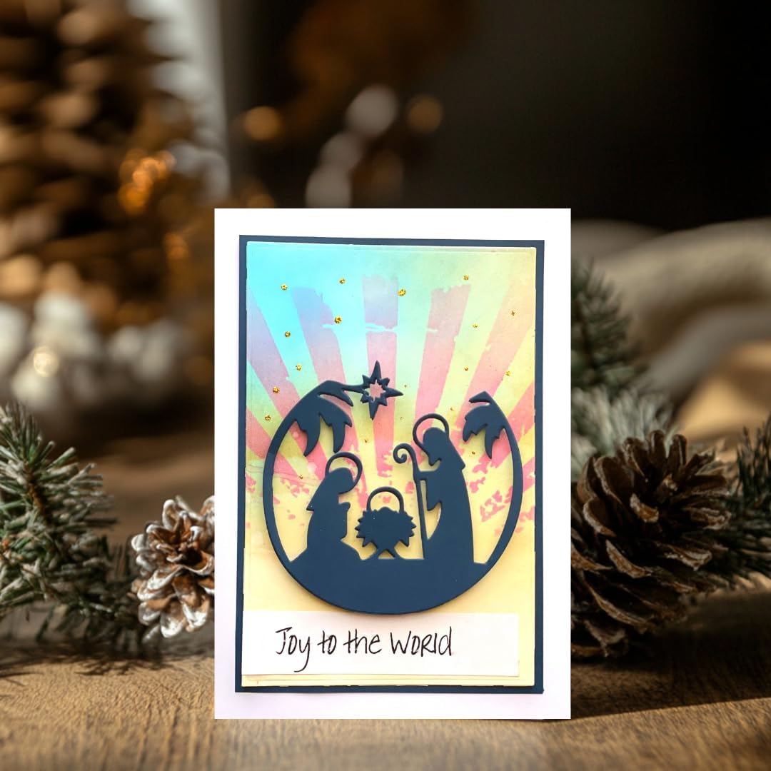 A handmade Christmas greeting card featuring a navy blue silhouette of the Holy Family and baby Jesus against a pastel rainbow sunburst background, with the text 'Joy to the World' at the bottom. The card is displayed on a wooden surface surrounded by pinecones and pine branches.