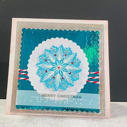 A handmade square Christmas greeting card with a layered design. The card features a light pink border, a teal blue background, a white scalloped circle containing a light blue and white snowflake, red and white striped paper accents, and a light blue banner at the bottom with 'MERRY CHRISTMAS' printed on it.
