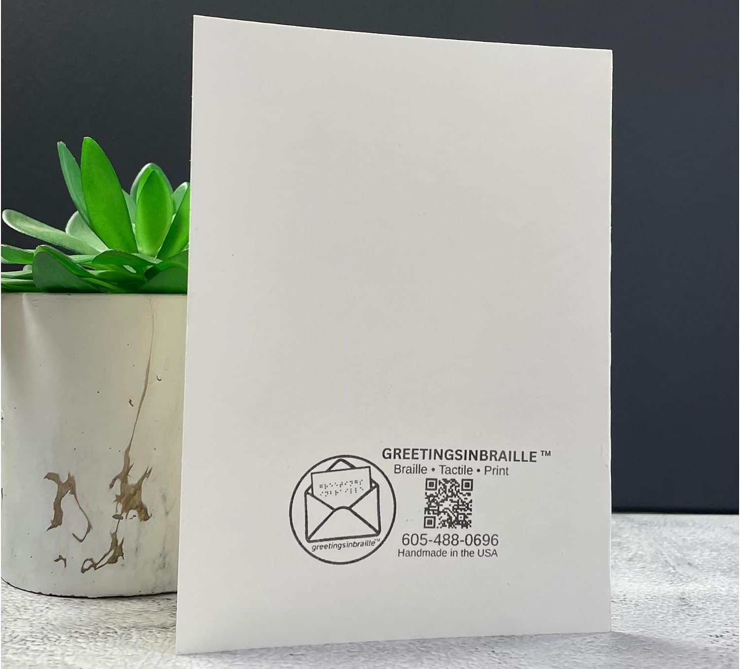 A white greeting card with black printed branding and a QR code, standing upright on a light surface next to a small green succulent in a white planter against a dark gray background.