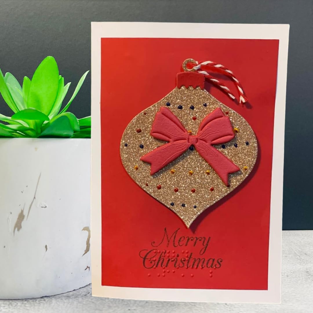 A handmade Christmas greeting card featuring a red background with a glittery gold ornament and a red bow, with 'Merry Christmas' written in black script. The card is styled next to a small potted succulent.