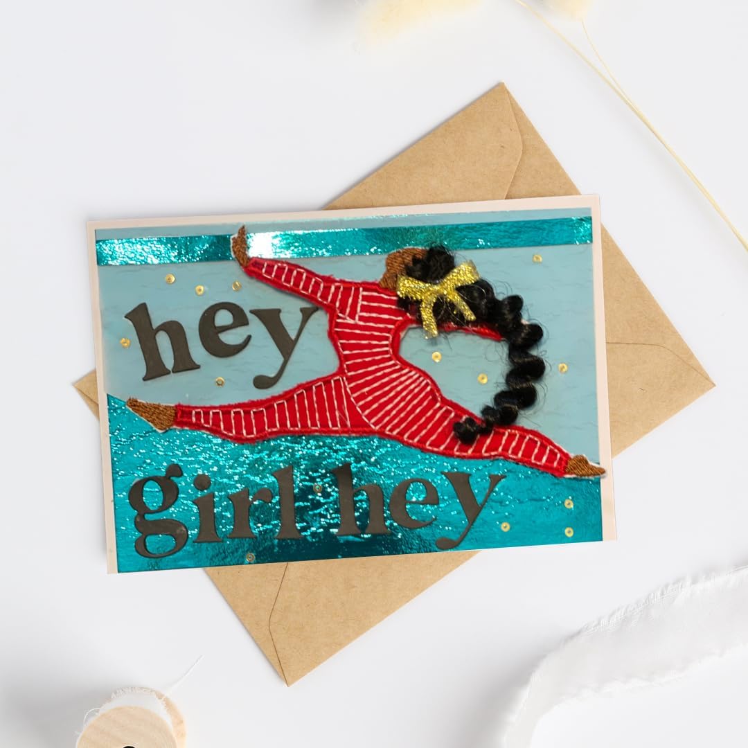 A handmade greeting card with a dancer figure made from red striped fabric, black pom-poms, and gold glitter, placed on top of a brown kraft envelope against a white background.