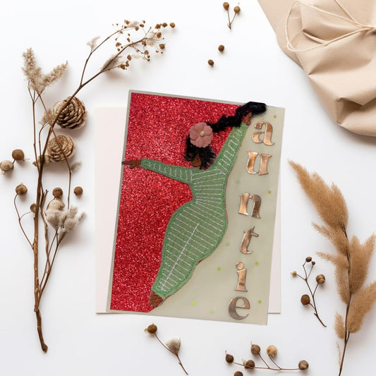 A handmade greeting card featuring a green dancing figure on a red glitter background, with the word 'auntie' spelled out in metallic letters, styled in a flat lay with dried botanicals and a beige fabric bow on a white surface.