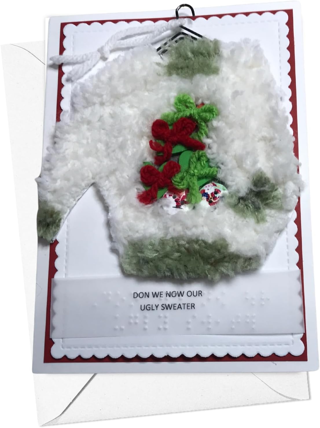 A handmade Christmas greeting card featuring a 3D fluffy white sweater ornament with green trim and red pomegranate embellishments. The card has a red border, white base, and the text 'DON WE NOW OUR UGLY SWEATER' with Braille below.