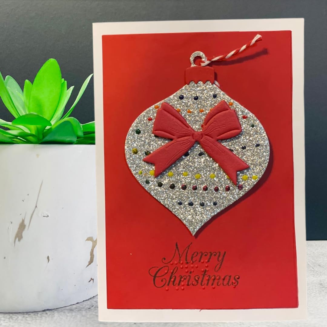 A handmade Christmas greeting card featuring a silver glitter ornament with a red bow on a red background, with 'Merry Christmas' written in black script. The card is styled next to a small potted succulent.