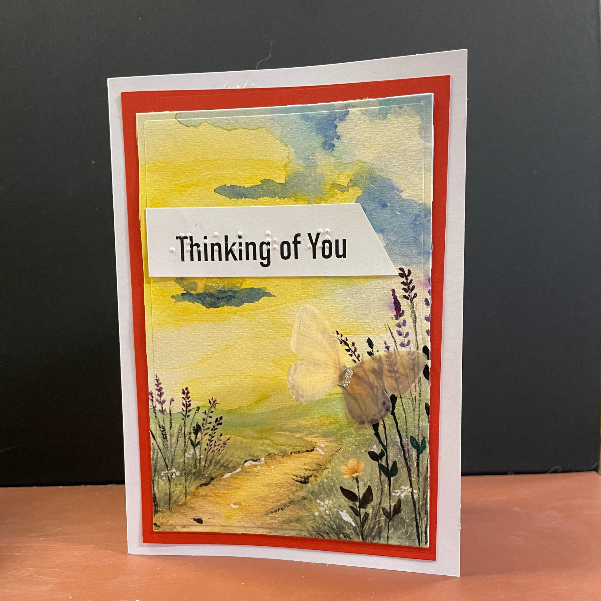 A handmade greeting card featuring a watercolor landscape of a path through wildflowers under a yellow and blue sky, with a white banner reading 'Thinking of You'. The card has a white border, a red inner border, and is standing on a wooden surface against a dark gray background.