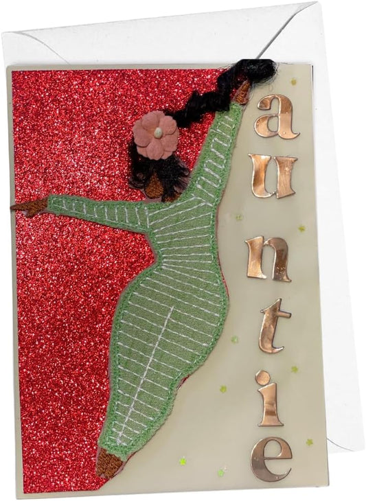A handmade greeting card featuring a stylized dancer silhouette in green and black with a pink flower, set against a red glitter background on an off-white card. The right side has metallic gold letters spelling 'auntie', and a white envelope is partially visible at the top.