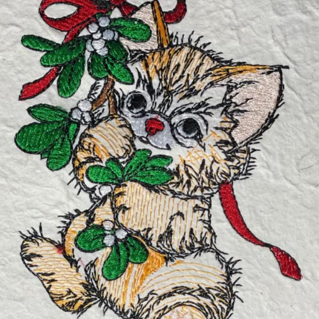 Close-up of a decorative embroidered design featuring a fluffy cream-colored kitten holding green mistletoe with red ribbons and white berries on a textured white fabric background.