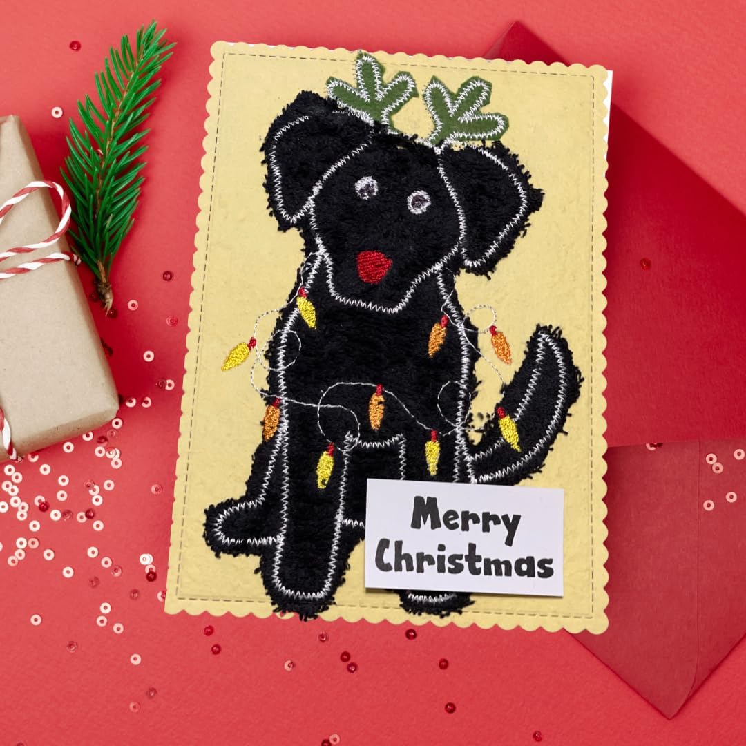 A handmade Christmas card featuring a black fabric dog decorated with green holiday leaves on its ears, colorful string lights, and a white 'Merry Christmas' label. The card is displayed on a red background with a gift and pine branch nearby.