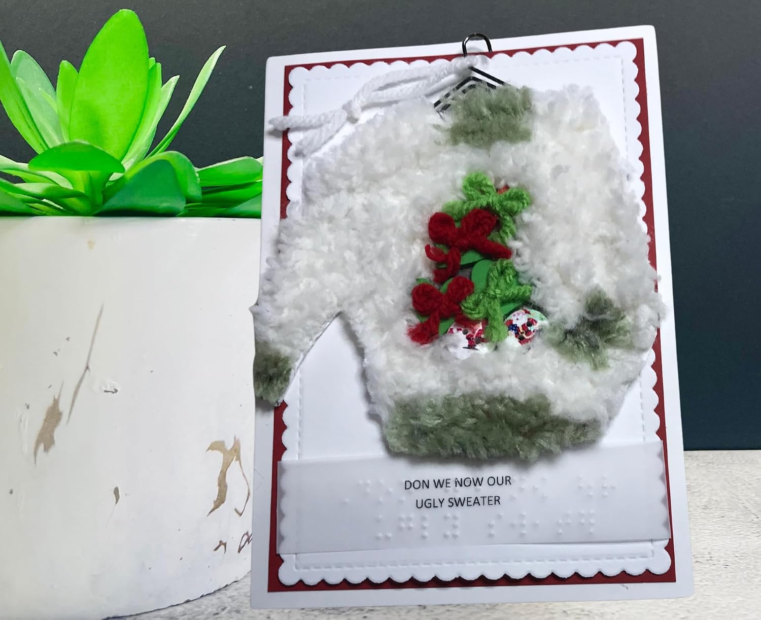 A handmade greeting card featuring a fuzzy white sweater with green and red patches, displayed next to a potted green plant against a dark gray background.
