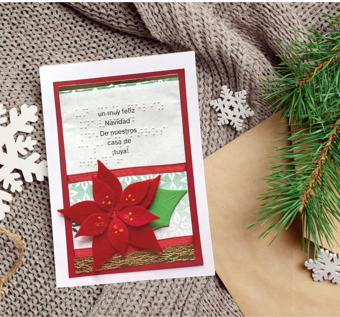 A handmade Christmas greeting card with Spanish text, featuring a red poinsettia and green leaf embellishment, styled on a brown knit blanket with pine branches, white snowflakes, and a kraft envelope.