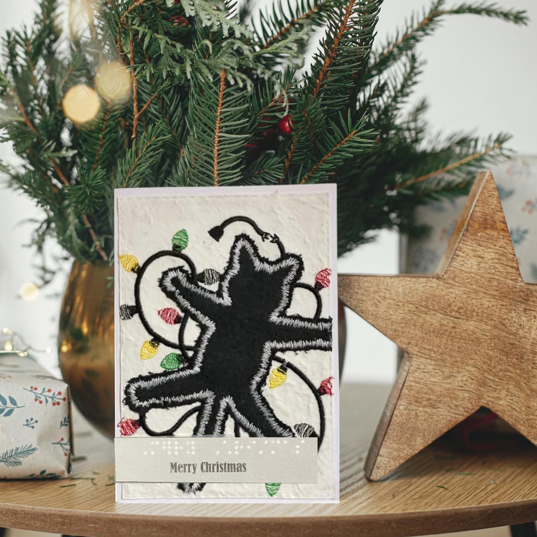 A handmade Christmas greeting card featuring a black embroidered star with colorful Christmas lights and the text 'Merry Christmas' at the bottom, styled on a wooden table with a pine tree and a wooden star ornament in the background.