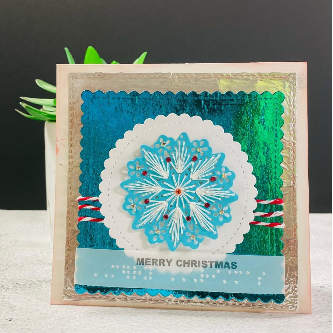 A handmade square Christmas greeting card with a blue and green gradient background, a white scalloped circle containing a blue snowflake design, and a light blue banner at the bottom that reads 'MERRY CHRISTMAS' with small white dots underneath. The card is displayed standing upright on a white surface with a dark gray background and a small potted plant behind it.