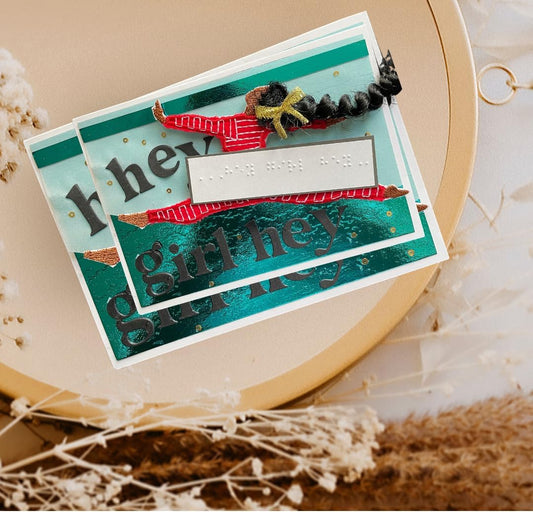 A handmade layered greeting card with 'girl hey' in black script over teal and red patterned paper, decorated with a red striped hat, gold foil dots, and a black curly hair embellishment, styled on a beige circular tray with dried flowers.