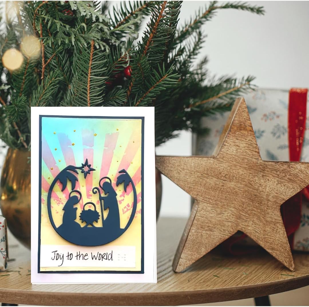 A handmade Christmas greeting card featuring a navy silhouette nativity scene on a pastel rainbow sunburst background, with the text 'Joy to the World' at the bottom. The card is styled on a wooden table next to a large wooden star and green pine branches.