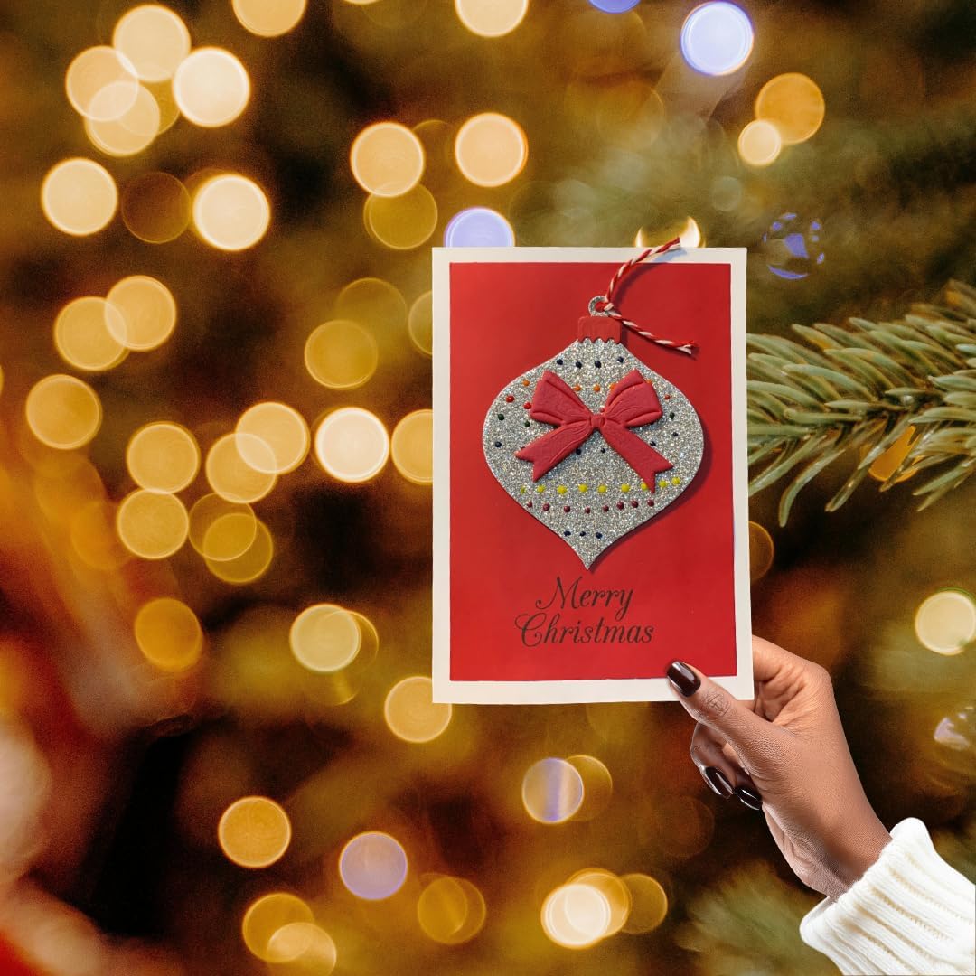 A hand holds a red Christmas greeting card featuring a glittery silver ornament with a red bow and the words 'Merry Christmas' against a blurred festive background of warm lights and pine branches.