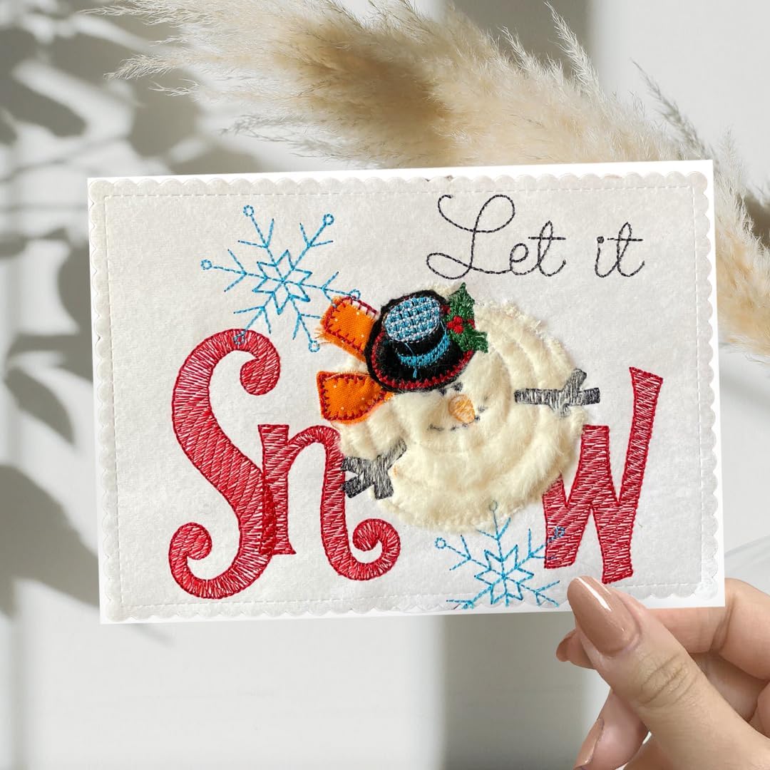 A handmade embroidered greeting card held by a hand, featuring the phrase 'Let it Snow' with a cream-colored snowman wearing a blue hat and surrounded by red and blue embroidery on a white fabric background.