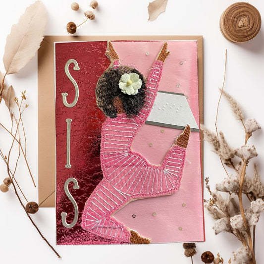 A handmade greeting card featuring a 3D cutout of a girl in a pink dance outfit with a white flower in her hair, set against a textured red and pink background with the letters 'SIS' in metallic silver. The card is displayed on a kraft envelope with dried botanicals on a white background.