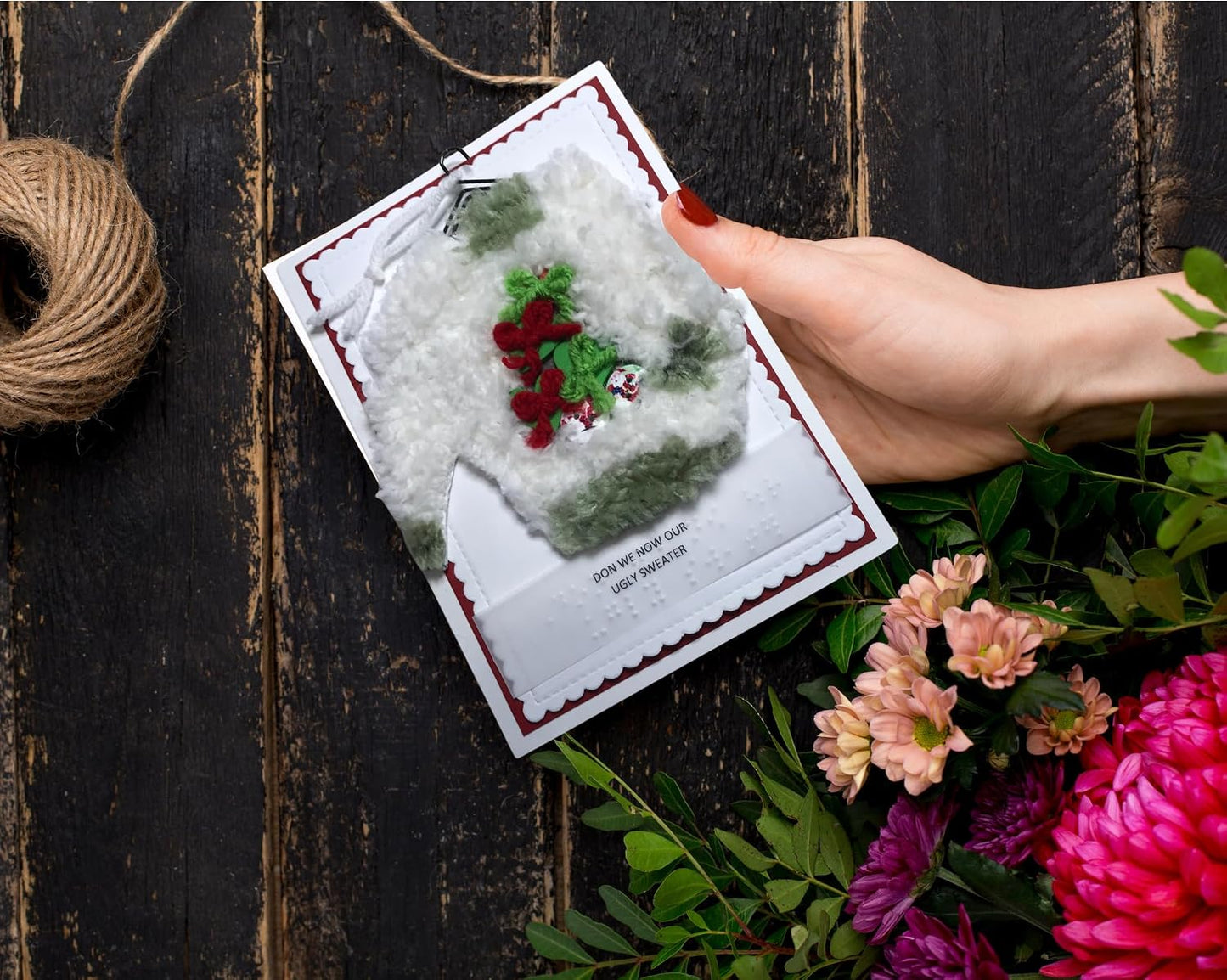 A hand holds a handmade greeting card featuring a white knit sweater with red and green Christmas embroidery, set against a dark rustic wooden background with flowers and twine.