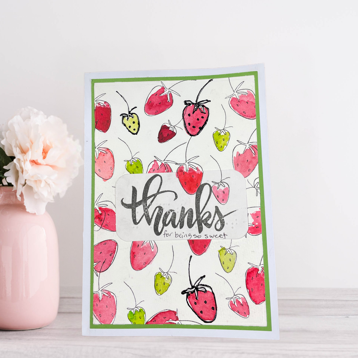 A handmade greeting card with a watercolor strawberry pattern and the word 'thanks' in script, styled on a light wood surface next to a pink vase with a white flower.