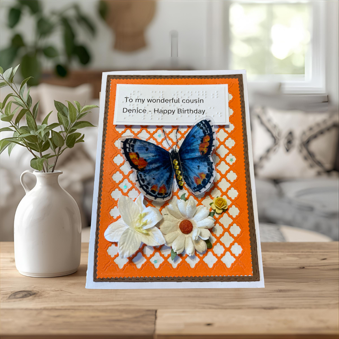 Greeting card with butterfly and flowers on an orange background, placed on a wooden surface with a vase and plant in the background.